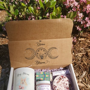 May include: A gift box with the text "The Divine Blossom" and a celestial design. Inside are a candle with a dog illustration and the words "Dog Mom", body butter, a bath bomb, lip balm, and matches.