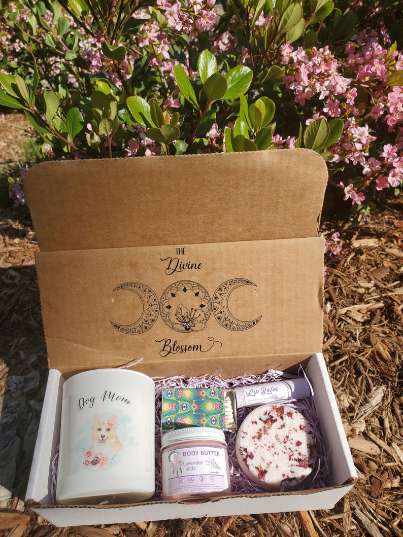 May include: A gift box with the text "The Divine Blossom" and a celestial design. Inside are a candle with a dog illustration and the words "Dog Mom", body butter, a bath bomb, lip balm, and matches.