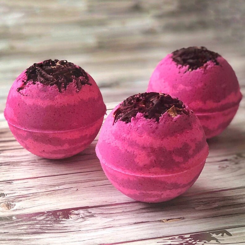 May include: Three pink bath bombs with a dark brown topping are arranged on a light wood surface. The bath bombs have a spherical shape with a textured surface. The dark brown topping is concentrated on the top of each bath bomb.