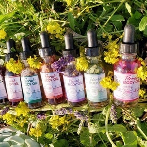 May include: A collection of seven glass dropper bottles filled with colorful floral body oils. Each bottle has a black dropper top and a label that reads "Floral Body Oil". The bottles are surrounded by green foliage and yellow flowers.