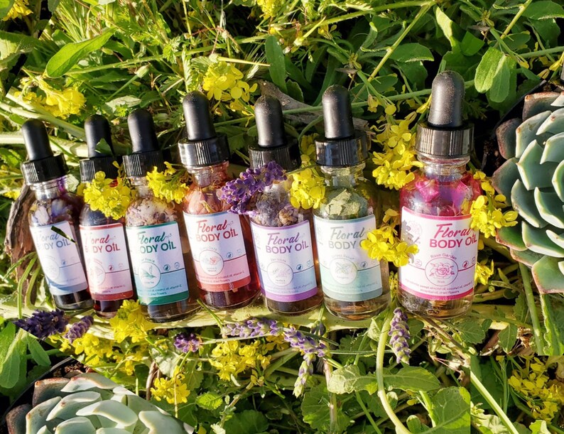 May include: A collection of seven glass dropper bottles filled with colorful floral body oils. Each bottle has a black dropper top and a label that reads "Floral Body Oil". The bottles are surrounded by green foliage and yellow flowers.