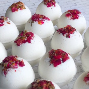 May include: A collection of white bath bombs, each topped with vibrant red and gold dried flower petals. The spherical bombs are arranged closely together, creating a visually appealing display. The overall aesthetic is clean and spa-like.