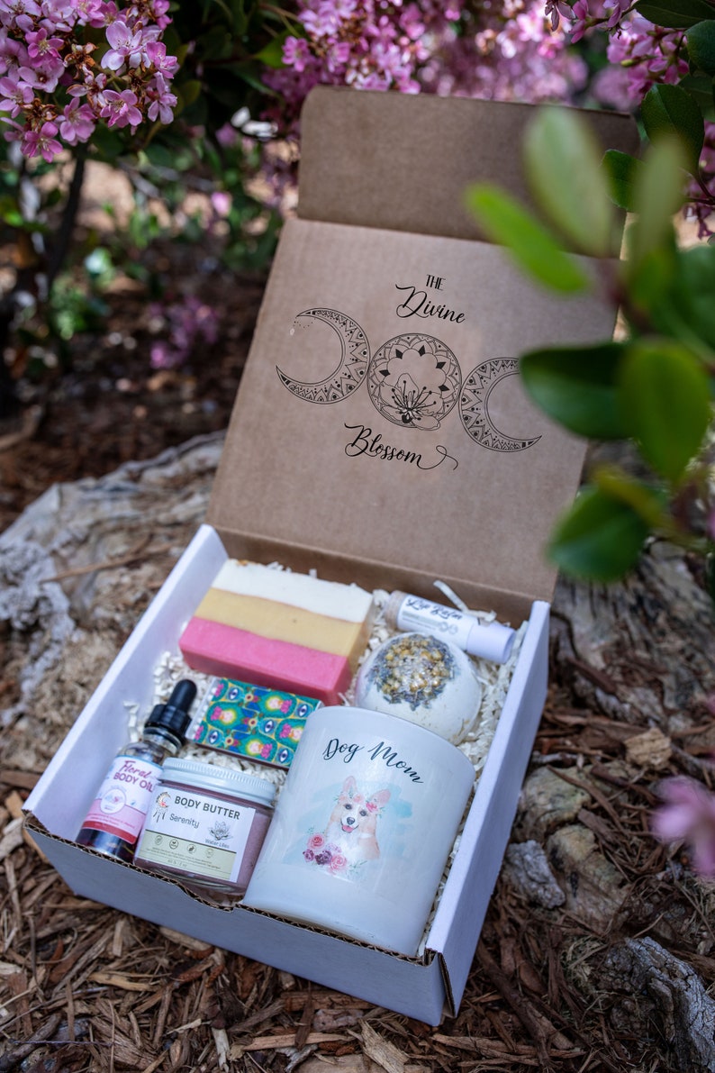 May include: A gift box with a variety of self-care items. The box includes a bar of soap, a bath bomb, a candle with a dog graphic, body oil, body butter, and a lip balm. The box is labeled "The Divine Blossom".