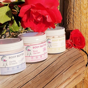 May include: Three jars of body butter with white lids and labels. The labels read "Vanilla Dreams", "Rose Garden", and "Lemon Kisses". The jars are arranged on a wooden surface, with red roses in the background. Each jar contains 2 fl oz of product.