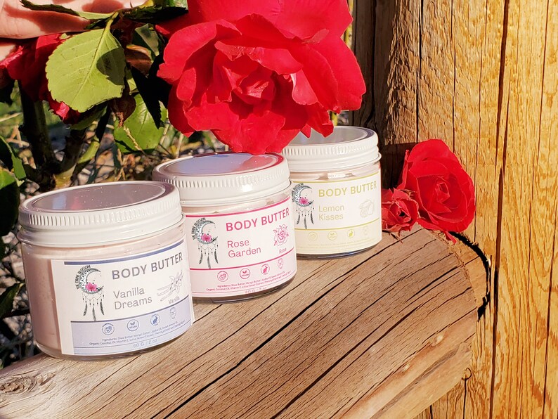 May include: Three jars of body butter with white lids and labels. The labels read "Vanilla Dreams", "Rose Garden", and "Lemon Kisses". The jars are arranged on a wooden surface, with red roses in the background. Each jar contains 2 fl oz of product.