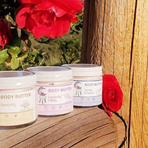 May include: Three glass jars of body butter with white lids are displayed on a wooden surface, with red roses in the background. Each jar has a label with the words "Body Butter" and a different scent name: Serenity, Lavender Fields, and Vanilla Dreams. Each jar contains 6 oz.