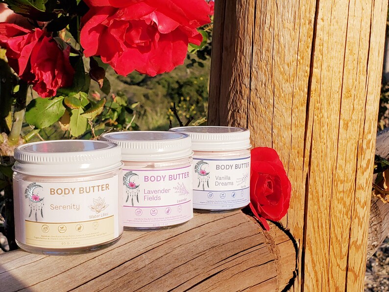 May include: Three glass jars of body butter with white lids are displayed on a wooden surface, with red roses in the background. Each jar has a label with the words "Body Butter" and a different scent name: Serenity, Lavender Fields, and Vanilla Dreams. Each jar contains 6 oz.
