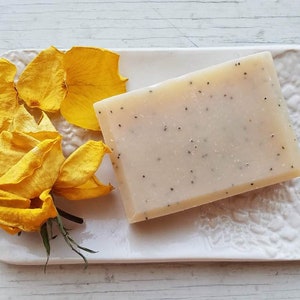 May include: A rectangular bar of handmade soap with small black seeds, resting on a white ceramic dish. Yellow rose petals and a rose are arranged beside the soap, creating a spa-like aesthetic. The soap is approximately 4 inches long.
