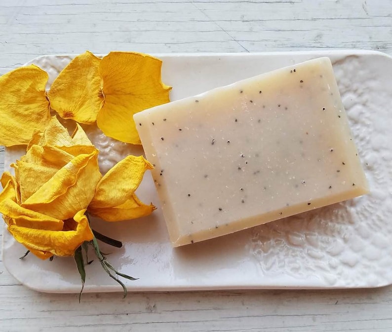 May include: A rectangular bar of handmade soap with small black seeds, resting on a white ceramic dish. Yellow rose petals and a rose are arranged beside the soap, creating a spa-like aesthetic. The soap is approximately 4 inches long.