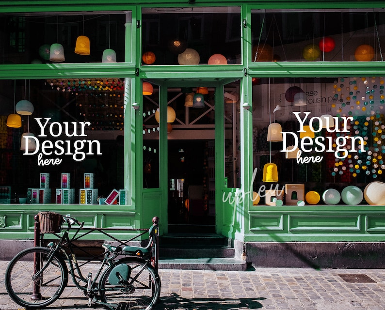 May include: A green storefront with two large windows displaying colorful decorations and the text "Your Design here". A bicycle is parked in front of the store.