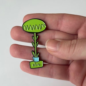 May include: Green enamel pin of a plant with a green pot and the word "Bleep" on the front.