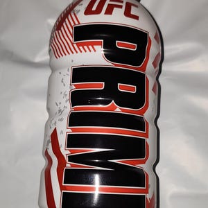 Może przedstawiać: Biała plastikowa butelka z czarną zakrętką i czerwono-czarnym logo PRIME. Butelka jest oznaczona jako "HYDRATION DRINK" i "16.9 FL OZ (500mL)". Butelka ma również logo UFC.