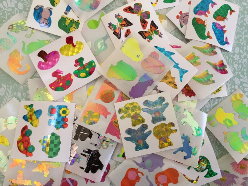 May include: A pile of holographic stickers with diverse designs, such as butterflies, animals, and abstract shapes. The stickers are on white backing paper and have a shiny, iridescent finish, creating a colourful and eye-catching effect.