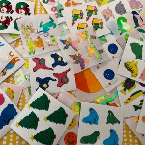 May include: A selection of colourful stickers with diverse designs, such as Santa Claus, Christmas trees, dogs, penguins, and musical notes. The stickers are on small, square sheets with a white background, scattered on a yellow dotted surface.