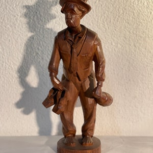 May include: A wooden carving of a man in traditional clothing holding a bottle and a pair of shoes. The carving is on a wooden base with the inscription "Der Heftpfeifer".