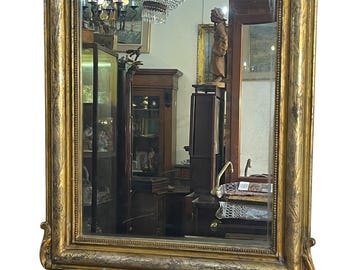 May include: An antique gold-framed mirror, rectangular with a rounded top and decorative feet. The frame features ornate carvings. The mirror reflects a room with antique furniture and a chandelier.