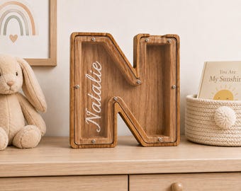 May include: A wooden letter N shaped piggy bank with a clear acrylic front panel secured by small metal screws. The name Natalie is printed in white script on the transparent surface, revealing the natural wood grain texture inside the frame.