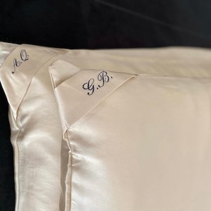 May include: Two white satin pillowcases with embroidered initials. The pillowcase on the left has the initials "A.Q." embroidered in blue thread. The pillowcase on the right has the initials "G.B." embroidered in blue thread.