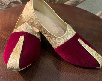 Op de afbeelding: Een paar schoenen van bordeauxrood fluweel met gouden borduurwerk. De schoenen hebben een spitse neus en een platte zool.