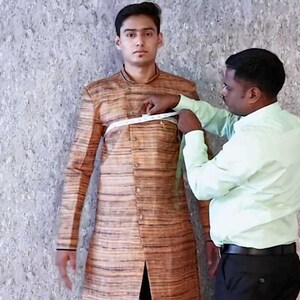 K&ouml;nnte beinhalten: Ein Schneider misst den Brustumfang einer Person, die eine braune und beige gestreifte Jacke tr&auml;gt. Der Schneider h&auml;lt ein wei&szlig;es Ma&szlig;band.