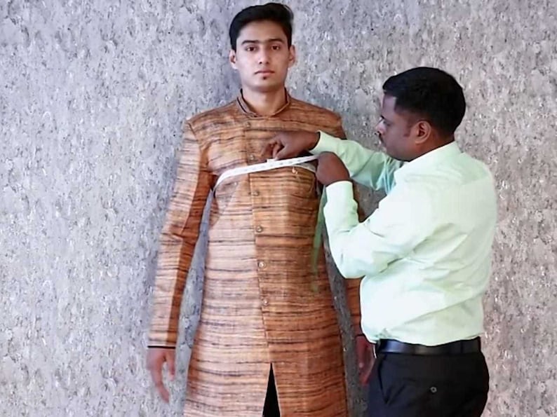 K&ouml;nnte beinhalten: Ein Schneider misst den Brustumfang einer Person, die eine braune und beige gestreifte Jacke tr&auml;gt. Der Schneider h&auml;lt ein wei&szlig;es Ma&szlig;band.