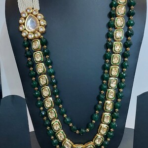 Pu&ograve; includere: Una collana a tre file con perline verdi e dettagli dorati. Ogni fila presenta ciondoli quadrati con pietre trasparenti incastonate in oro. La collana ha un ciondolo grande e decorato con una pietra trasparente al centro.