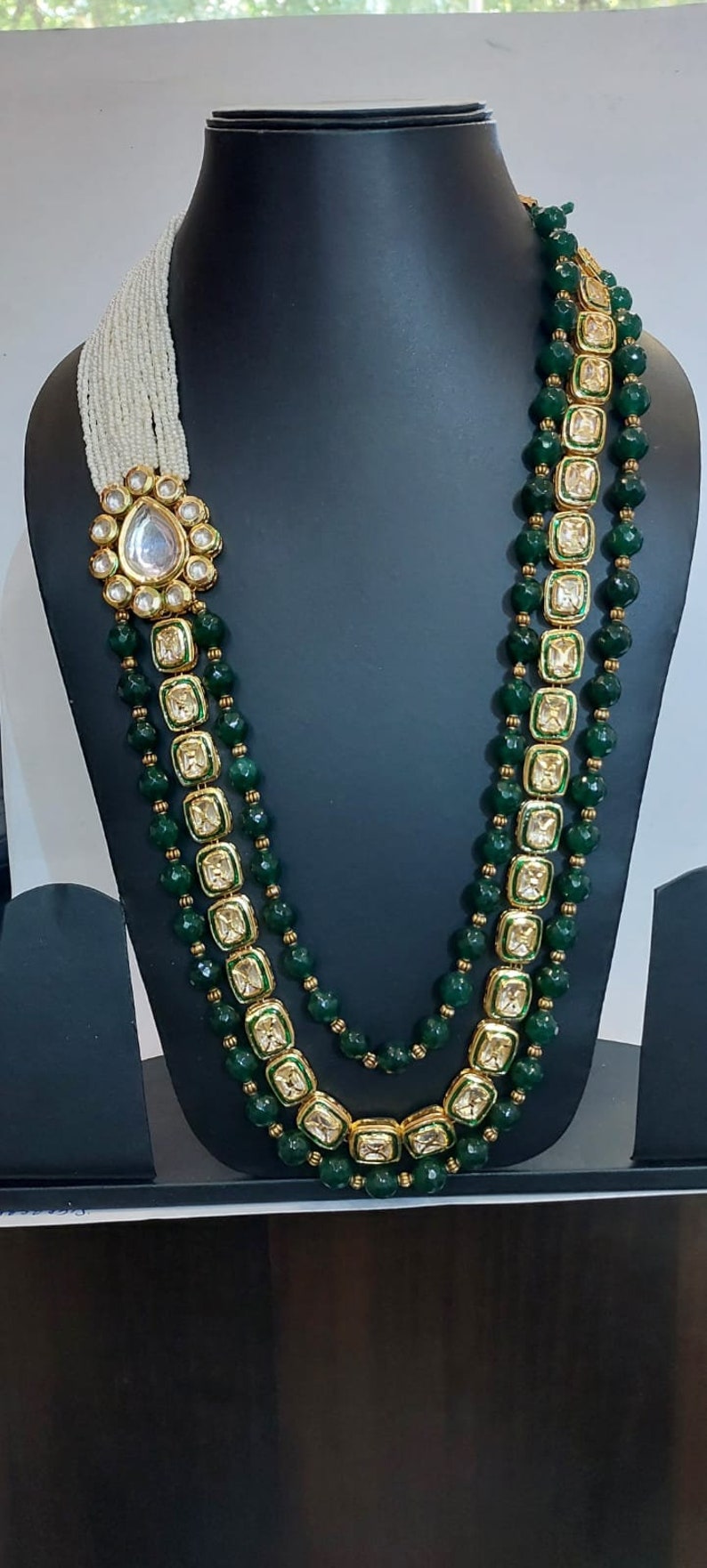 Pu&ograve; includere: Una collana a tre file con perline verdi e dettagli dorati. Ogni fila presenta ciondoli quadrati con pietre trasparenti incastonate in oro. La collana ha un ciondolo grande e decorato con una pietra trasparente al centro.