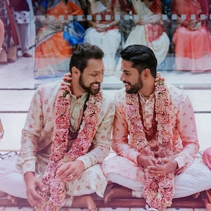 Può includere: Due persone vestite con abiti tradizionali indiani, con ghirlande di fiori intorno al collo, sono sedute e si sorridono.