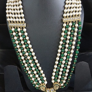 Pu&ograve; includere: Una collana multifilo con perline bianche e verdi. La collana ha un ciondolo dorato con un design floreale e un fiocco.
