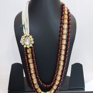 Pu&ograve; includere: Una collana multifilo con perline color borgogna e dettagli dorati. La collana presenta un grande ciondolo decorato con una pietra trasparente al centro.