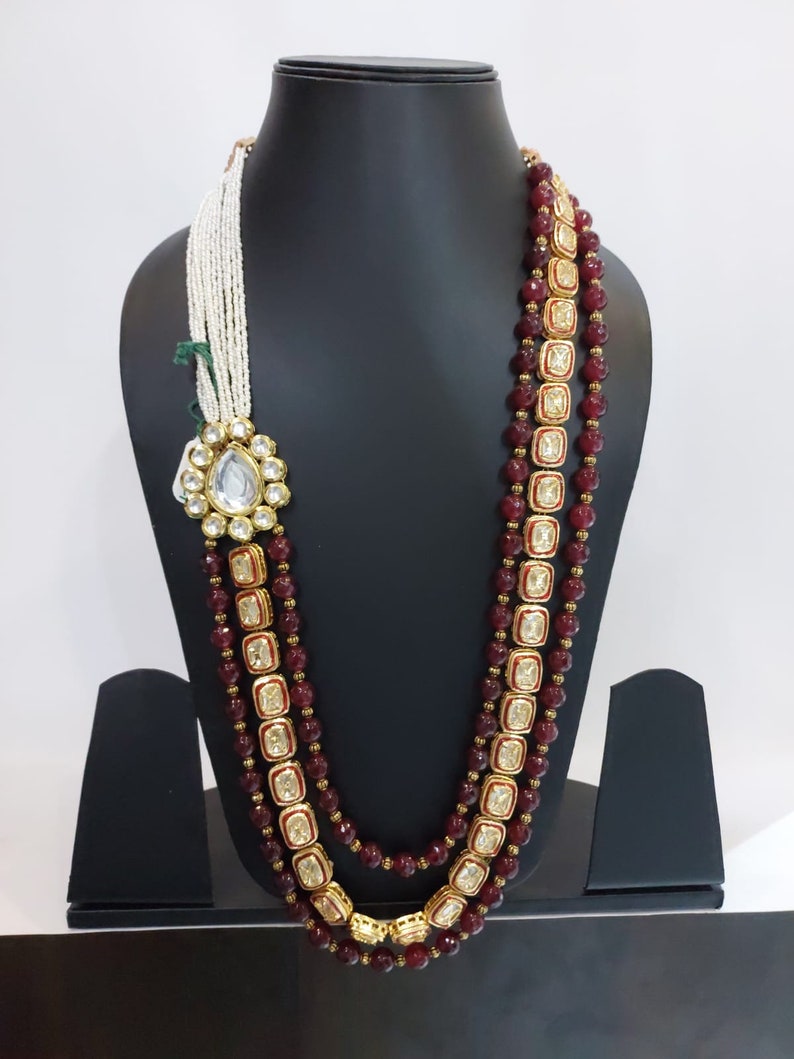 Pu&ograve; includere: Una collana multifilo con perline color borgogna e dettagli dorati. La collana presenta un grande ciondolo decorato con una pietra trasparente al centro.