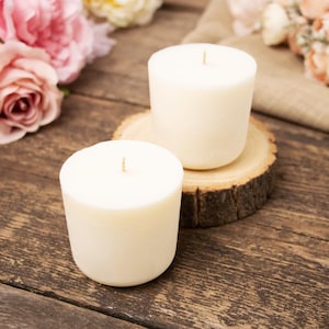 May include: Two cylindrical off-white votive candles with a single centred wick each. One candle sits directly on a dark wooden surface, while the other is placed on a circular wooden slice with visible bark edges, surrounded by pink flowers.