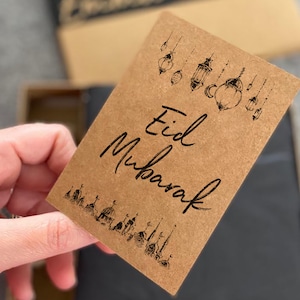 May include: A brown card with the text "Eid Mubarak" written in black calligraphy. The card is decorated with black illustrations of lanterns and mosques.
