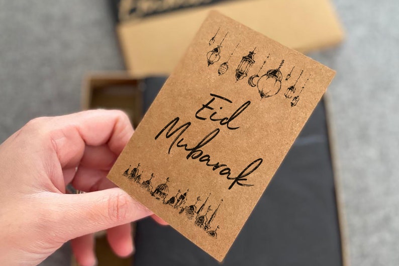 May include: A brown card with the text "Eid Mubarak" written in black calligraphy. The card is decorated with black illustrations of lanterns and mosques.