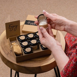 May include: A brown box with a set of small glass jars filled with candles. The box has the words "Enjoy" and "Happy" printed on it. The jars are arranged in a grid pattern and have black lids. A small burlap bag is also included in the box.