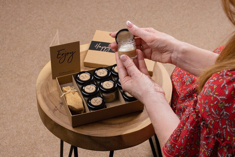 May include: A brown box with a set of small glass jars filled with candles. The box has the words "Enjoy" and "Happy" printed on it. The jars are arranged in a grid pattern and have black lids. A small burlap bag is also included in the box.