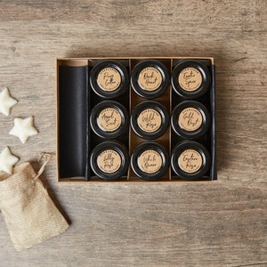 May include: A set of nine small glass jars with black lids and brown paper labels. The jars are arranged in a 3x3 grid in a wooden box. The labels have different names of rose scents, such as "Pink Cotton", "Wild Rose", and "Eastern Rose".