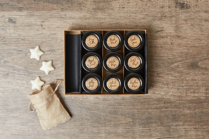 May include: A set of nine small glass jars with black lids and brown paper labels. The jars are arranged in a 3x3 grid in a wooden box. The labels have different names of rose scents, such as "Pink Cotton", "Wild Rose", and "Eastern Rose".