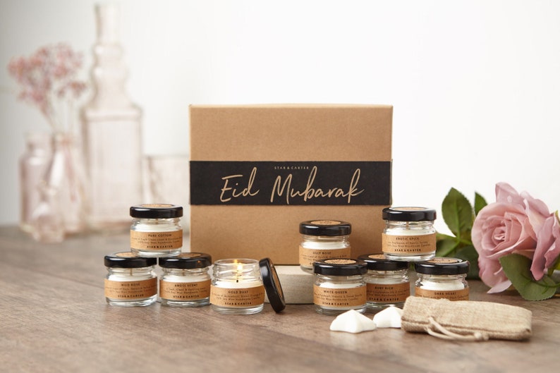 May include: A set of 10 small glass candle jars with black lids, arranged in a circle around a brown box with a black ribbon that says "Eid Mubarak". The candles are white and have a variety of scents, including "Wild Rose", "White Cotton", and "Angel Glow".