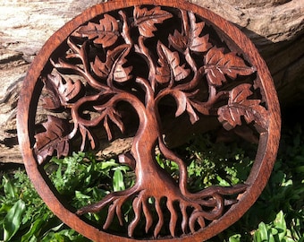 May include: A round wooden wall hanging with a carved tree of life design. The tree has intricate branches and leaves, and the roots are deeply intertwined. The wood is a rich brown color.