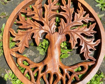 May include: A round, wooden wall hanging featuring a detailed carving of a tree of life. The tree has a trunk, roots, and leafy branches, all within a circular frame. The wood has a warm, brown tone, and the carving is intricate.
