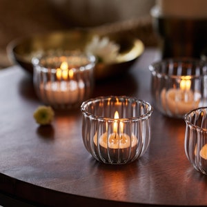 Pode incluir: Quatro porta-velas de vidro com lados canelados, cada um com uma vela de chá acesa. Os porta-velas estão sobre uma mesa de madeira escura.