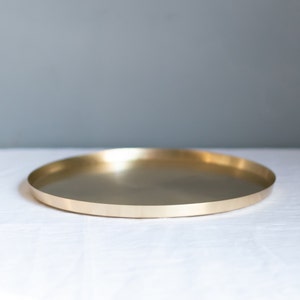 May include: A round, gold-colored metal tray with a smooth, reflective surface. The tray is sitting on a white surface.