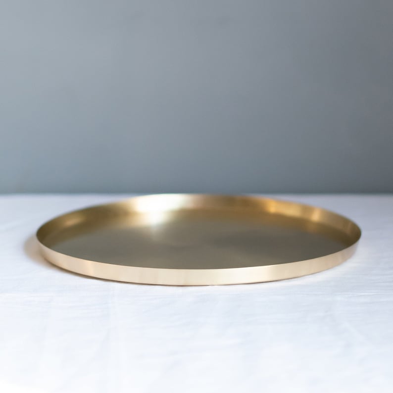 May include: A round, gold-colored metal tray with a smooth, reflective surface. The tray is sitting on a white surface.