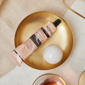 May include: A gold metal tray with a tube of Aesop Resurrection Aromatique Hand Balm and a white marble sphere. The tube is labeled "Resurrection Aromatique Hand Balm" and "Aesop".