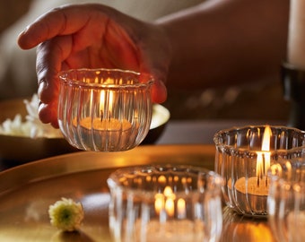 May include: Four clear glass candle holders with fluted sides, each holding a white candle. The candles are lit and casting a warm glow. The candle holders are arranged on a gold tray.