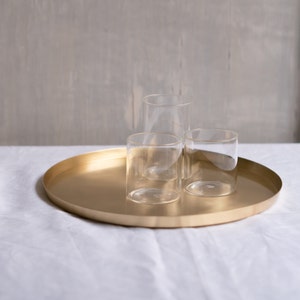 May include: Three clear glass tumblers on a gold round tray. The tray is on a white linen tablecloth.