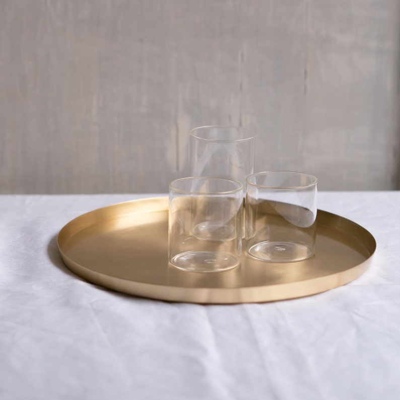 May include: Three clear glass tumblers on a gold round tray. The tray is on a white linen tablecloth.
