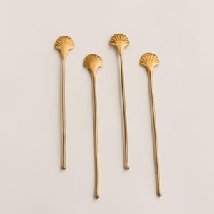 May include: A set of four gold-toned metal stirrers, each featuring a distinct fan-shaped shell design at the top and a slender, elongated stem that terminates in a small, rounded tip at the bottom, arranged vertically against a plain background.