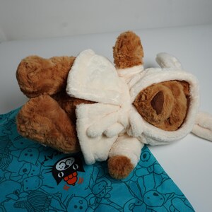 May include: A brown plush teddy bear wearing a cream-colored hooded robe with long ears attached to the hood. The bear is positioned on top of a teal fabric bag featuring a repeating pattern of black line-art cat faces and the word CAT.
